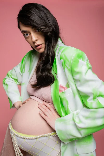 Pregnant woman with wavy brunette hair, wearing green and white blazer, crop top and beads belt, embracing tummy and looking at camera isolated on pink, stylish maternity concept — Stock Photo