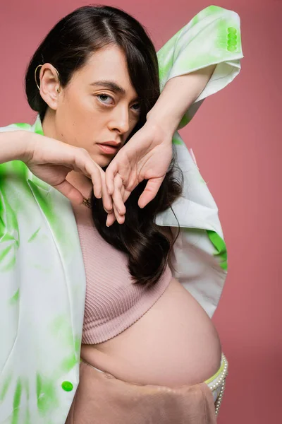 Donna incinta alla moda in crop top con blazer verde e bianco che si tiene per mano vicino al viso e guardando la fotocamera isolata su sfondo rosa, concetto di stile maternità — Foto stock