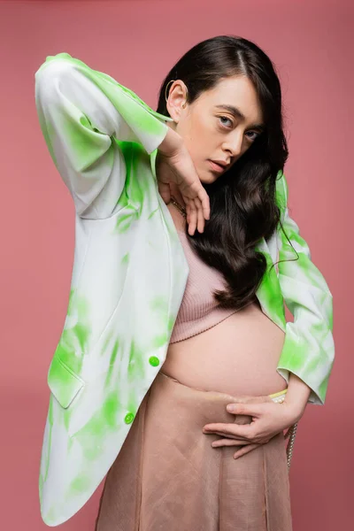 Brunette mother-to-be holding hand near face and looking at camera while posing in green and white blazer and crop top isolated on pink background, maternity fashion concept — Stock Photo