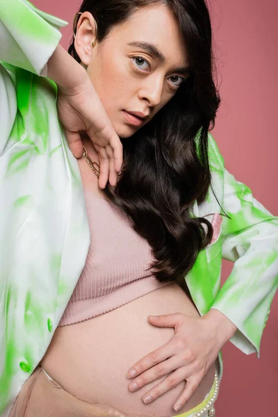 Charmante femme enceinte avec des cheveux bruns ondulés posant dans le haut de la culture et blazer vert et blanc tout en regardant la caméra isolée sur fond rose, concept de maternité à la mode — Photo de stock
