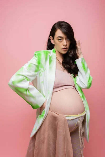 Attraente futura madre in blazer verde e bianco, crop top e leggings con cintura di perline e panno di chiffon guardando la fotocamera isolata su sfondo rosa, elegante concetto di gravidanza — Foto stock