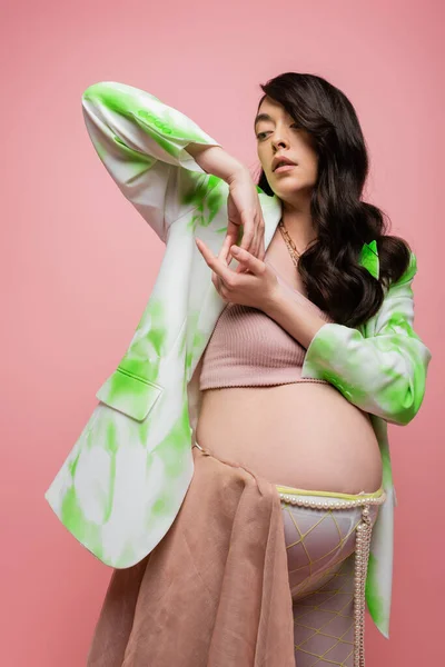 Fashionable future mother with wavy brunette hair posing in green and white blazer, crop top and beads belt with chiffon cloth isolated on pink background, pregnancy style concept — Stock Photo