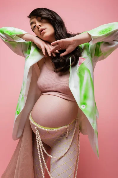 Brunette pregnant woman in green and white blazer, crop top, leggings with beads belt and chiffon cloth holding hands under chin and looking at camera isolated on pink, maternity fashion concept — Stock Photo