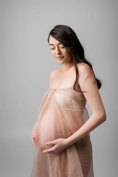 Очаровательная будущая брюнетка в воздушной ткани шифона и золотых браслетах касаясь живота и улыбаясь изолированы на сером фоне, концепция моды материнства, беременная женщина — стоковое фото