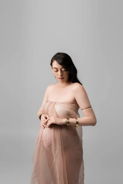 Витончена мати-бути з хвилястим волоссям брюнетки, позує в золотих браслетах і м'який шифон драпірування ізольовані на сірому фоні, концепція моди материнства, вагітна жінка — стокове фото