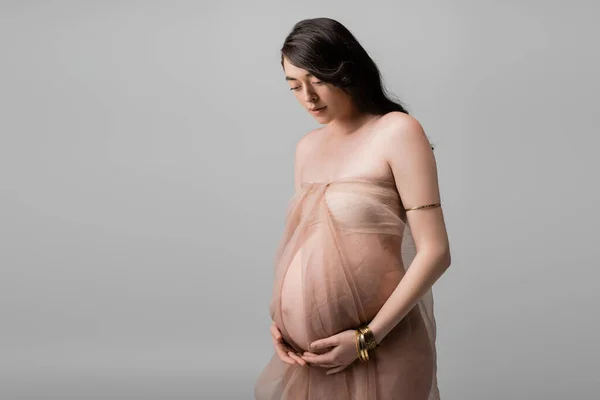 Элегантная ожидающая мать с волнистыми волосами брюнетки, стоящая в бежевой шифон ткани и золотые браслеты изолированы на сером фоне, концепция моды материнства, беременная женщина — стоковое фото