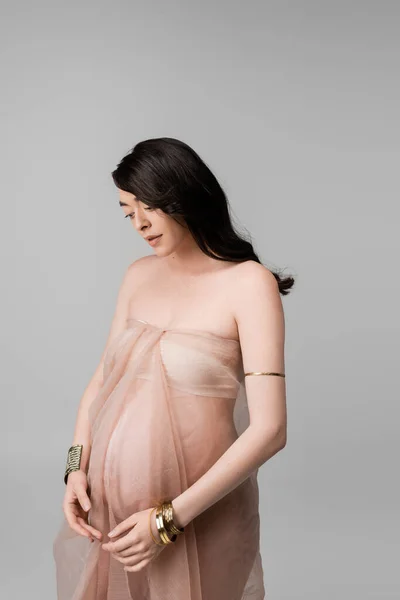Anmutige schwangere Frau mit welligen brünetten Haaren posiert mit beigem Chiffontuch und goldenen Armbändern auf grauem Hintergrund, Umstandsmodekonzept, Erwartung — Stockfoto