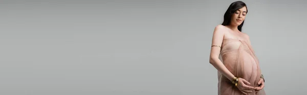 Femme enceinte romantique et élégante en mousseline de soie aérée drapé et accessoires dorés debout isolé sur fond gris, concept de mode maternité, bannière, sensualité — Photo de stock
