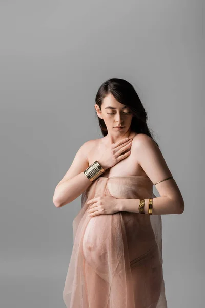 Brunette pregnant woman in golden accessories touching chest while posing in gentle chiffon draping isolated on grey background, maternity style concept, sensuality — Stock Photo