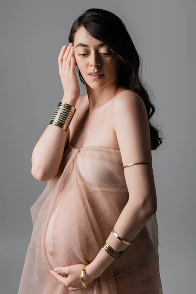 Romántica madre embarazada en pulseras de oro y suave gasa drapeado de la mano cerca de la cara aislada sobre fondo gris, concepto de moda de maternidad, sensualidad - foto de stock