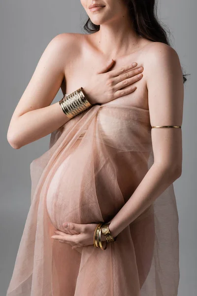 Cropped view of stylish and elegant pregnant woman posing in golden accessories and transparent chiffon draping isolated on grey background, maternity fashion concept, future mother with belly — Stock Photo