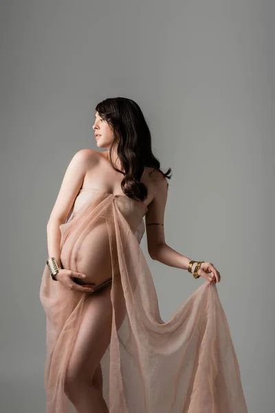 Graceful and pregnant woman with wavy brunette hair posing in transparent chiffon draping and touching belly isolated on grey background, maternity fashion concept, future mother with belly — Stock Photo