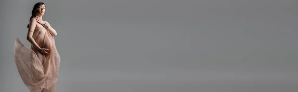 Donna incinta con capelli castani ondulati in posa in bracciali dorati e tessuto chiffon trasparente e fluente isolato su sfondo grigio, concetto di moda maternità, banner — Foto stock