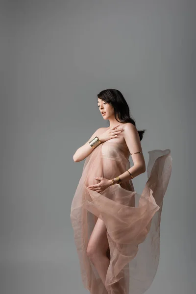 Romantic mother-to-be touching chest while posing in golden bracelets and delicate chiffon draping isolated on grey background, maternity fashion concept, pregnant woman with belly — Stock Photo