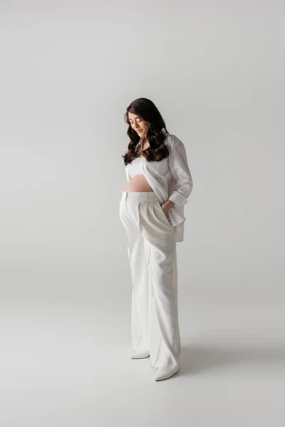 Full length of brunette pregnant woman in white shirt and crop top standing with hand in pocket of pants on grey background, fashionable pregnancy concept, expectation, future mother with belly — Stock Photo