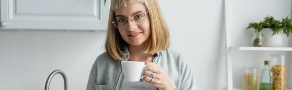 Счастливая молодая женщина с короткими волосами и челкой и очками, держа чашку утреннего кофе, стоя в повседневной одежде рядом с кухонным шкафом и растением в современной квартире, баннер — стоковое фото
