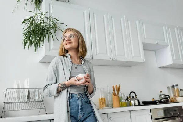 Joyful young woman with short hair and bangs, eyeglasses and tattoo holding cup of morning coffee while looking away and standing in casual clothes next to dishes, kettle, kitchen appliances — Stock Photo