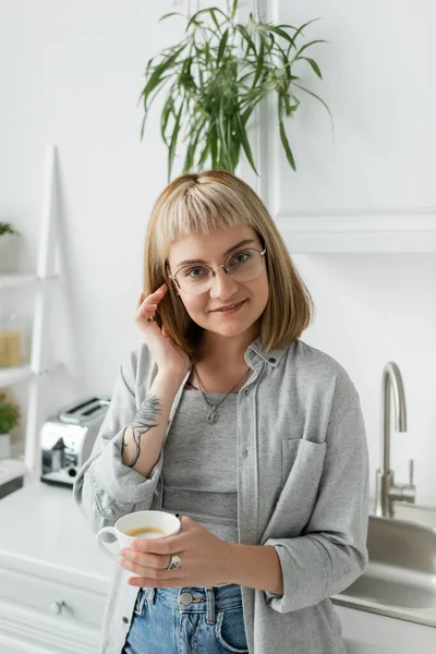 Счастливая молодая женщина с челкой, очками и татуировкой на руке регулируя короткие волосы и держа чашку кофе, глядя в камеру, стоя в повседневной одежде рядом с тостером, кухонная раковина дома — стоковое фото