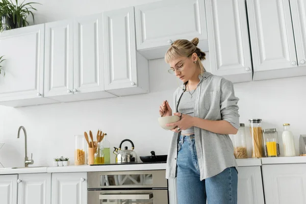 Young woman with bangs, eyeglasses and short hair holding bowl with breakfast and spoon while standing in casual grey clothes next to kettle, kitchen appliances and blurred white cabinets at home — Stock Photo