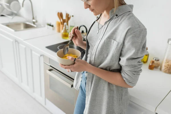 Vista cortada de jovem mulher em óculos segurando tigela com flocos de milho e colher, enquanto em pé em roupas cinzentas casuais ao lado de aparelhos de cozinha em cozinha branca borrada em casa — Fotografia de Stock