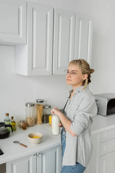 Mujer joven en gafas graduadas y pelo corto con flequillo sosteniendo botella con leche cerca de tazón con copos de maíz mientras hace el desayuno y de pie en ropa casual al lado de electrodomésticos de cocina en casa - foto de stock