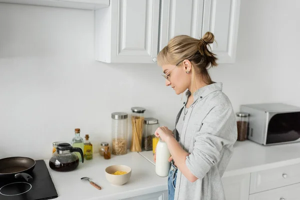 Mujer joven en gafas graduadas y pelo corto con flequillo sosteniendo botella con leche fresca cerca de un tazón con copos de maíz mientras hace el desayuno y de pie en ropa casual al lado de los electrodomésticos de cocina en casa - foto de stock
