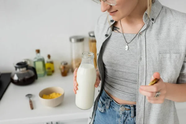 Обрезанный вид татуированной женщины, держащей бутылку со свежим молоком возле размытой чаши с кукурузными хлопьями и ложкой во время завтрака и стоя в повседневной одежде рядом с кухонной техникой дома — стоковое фото
