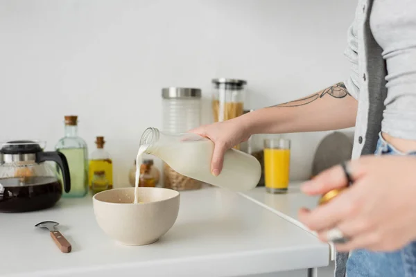 Vista recortada de mujer joven tatuada sosteniendo botella mientras vierte leche fresca en un tazón con copos de maíz en la encimera de la cocina mientras hace el desayuno y de pie en ropa casual en la cocina moderna - foto de stock