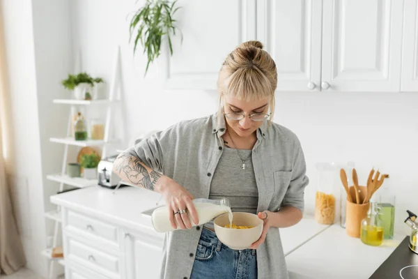 Mujer joven tatuada con flequillo en gafas con botella mientras vierte leche fresca en un tazón con copos de maíz y hace el desayuno mientras está de pie en ropa casual en la cocina moderna - foto de stock