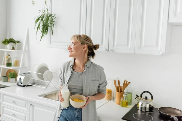 Tattooed young woman with bangs in eyeglasses holding bottle with fresh milk and bowl with cornflakes while making breakfast and looking away, standing in casual clothes in modern kitchen — Stock Photo