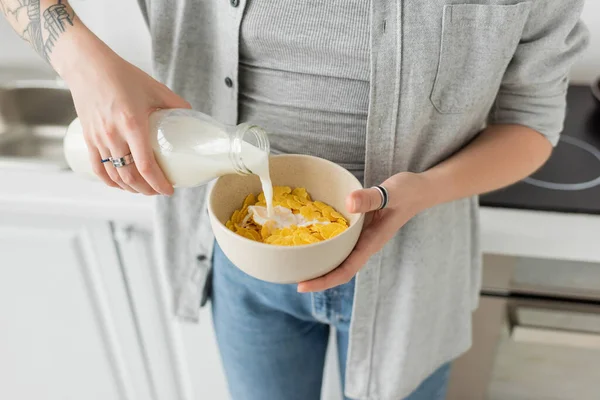 Cropped view of tattooed young woman holding bottle while pouring fresh milk into bowl with cornflakes and making breakfast while standing in casual clothes with denim jeans in modern kitchen — Stock Photo