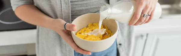 Cropped view of tattooed young woman holding bottle while pouring fresh milk into bowl with cornflakes and making breakfast while standing in casual clothes with denim jeans in modern kitchen, banner — Stock Photo