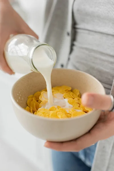Close up view of young woman holding bottle while pouring fresh milk into bowl with cornflakes and making breakfast while standing in casual clothes in modern kitchen — Stock Photo