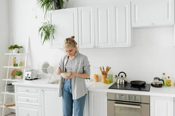 Молодая женщина с челкой в очках, держа миску с кукурузными хлопьями и ложкой, стоя в повседневной серой одежде и джинсах рядом с кухонной техникой в размытой белой кухне дома — стоковое фото