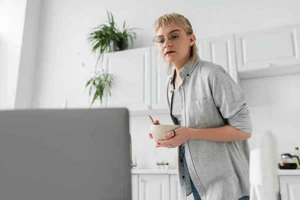 Curious young freelancer with bangs and eyeglasses holding bowl with cornflakes while looking at blurred laptop in modern kitchen with white cabinets and green plants on blurred background — Stock Photo