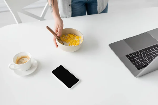 Cropped view of young freelancer with tattoo on hand holding bowl with cornflakes and spoon near laptop, smartphone with blank screen and cup of coffee on white desk in modern kitchen — Stock Photo