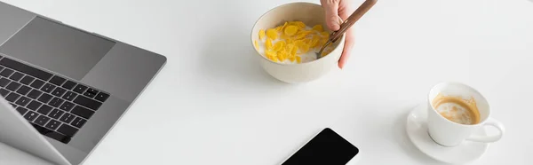 Vista recortada de un joven freelancer sosteniendo un tazón con hojuelas de maíz y cuchara cerca de un teléfono inteligente portátil con pantalla en blanco y una taza de café en un escritorio blanco en la cocina moderna, pancarta - foto de stock