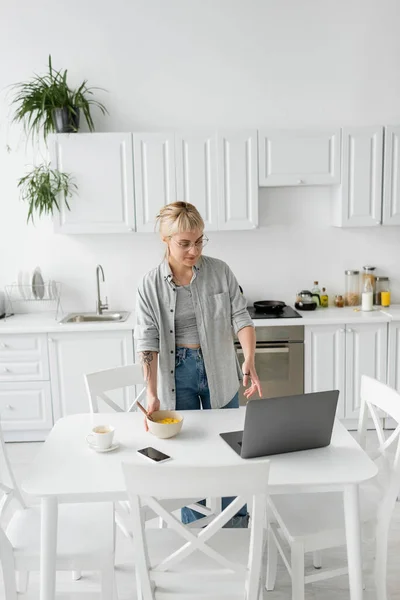 Tattooed freelancer with bangs and eyeglasses holding bowl with cornflakes while having breakfast and looking at laptop near smartphone and cup of coffee on saucer and desk in modern kitchen — Stock Photo