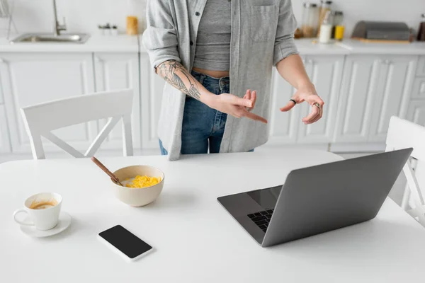 Cropped view of young freelancer with tattoo on hand pointing at laptop near smartphone with blank screen, bowl with cornflakes, spoon and cup of coffee on white saucer on desk in modern kitchen — Stock Photo