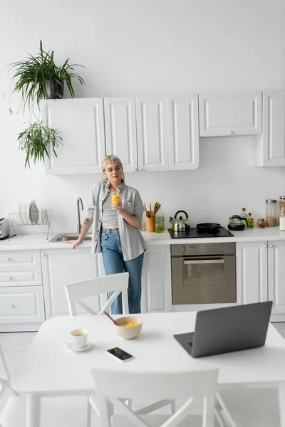 Tattooed woman in eyeglasses holding glass of orange juice and standing near desk with devices, bowl with cornflakes and cup of coffee with saucer on desk in modern kitchen, looking at laptop — Stock Photo