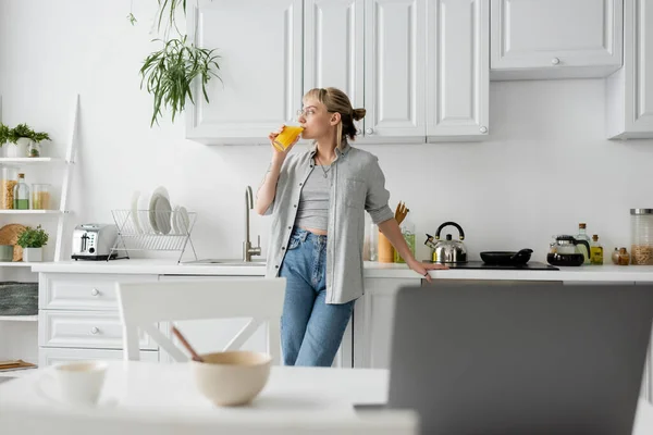 Mulher tatuada em óculos bebendo suco de laranja e de pé perto da bancada da cozinha ao lado da mesa com dispositivos, tigela com flocos de milho e xícara de café com pires em casa, estilo de vida freelance — Fotografia de Stock