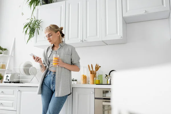 Tattooed woman with bangs and eyeglasses holding glass of orange juice and using smartphone while standing near clean dishes and green plants in blurred white kitchen in modern apartment — Stock Photo