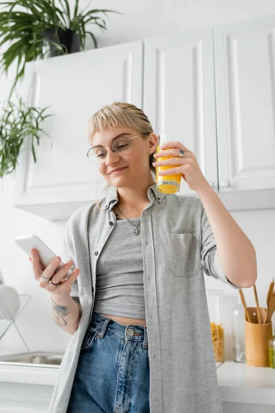 Mulher tatuada e feliz com franja e óculos segurando vidro de suco de laranja e usando smartphone enquanto está perto de pratos limpos e plantas verdes desfocadas no apartamento moderno — Fotografia de Stock