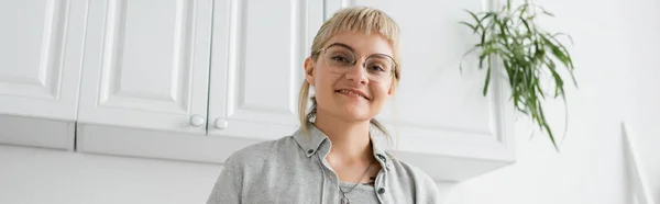 Happy young woman with bangs and eyeglasses smiling while standing in grey casual outfit and looking at camera near white kitchen cabinets and blurred green plants in modern apartment, banner — Stock Photo