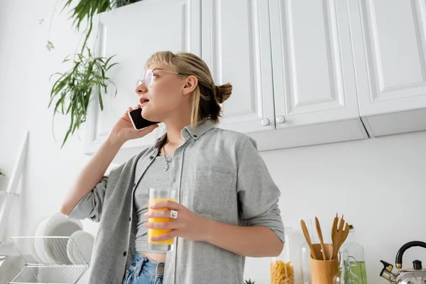 Low angle view of young woman with bangs and eyeglasses holding glass of orange juice and talking on smartphone while looking away and standing in kitchen and blurred green plants in modern apartment — Stock Photo