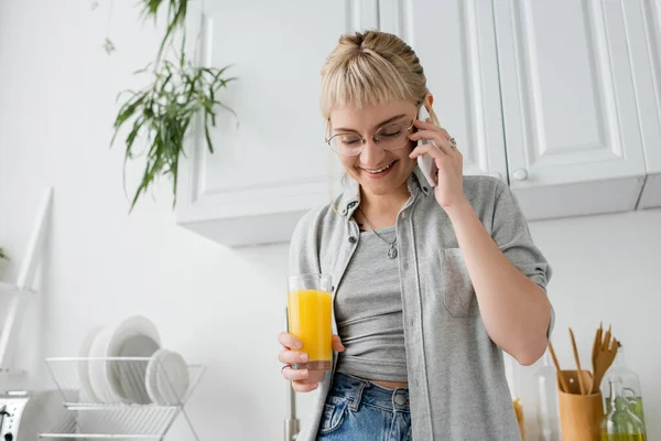 Happy young woman with bangs and eyeglasses holding glass of orange juice and talking on smartphone standing with closed eyes in kitchen near blurred green plants in modern apartment — Stock Photo