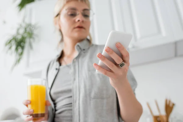 Young woman in eyeglasses, with ring on finger holding glass of orange juice and smartphone while texting and standing in blurred white kitchen with green indoor plants in modern apartment — Stock Photo