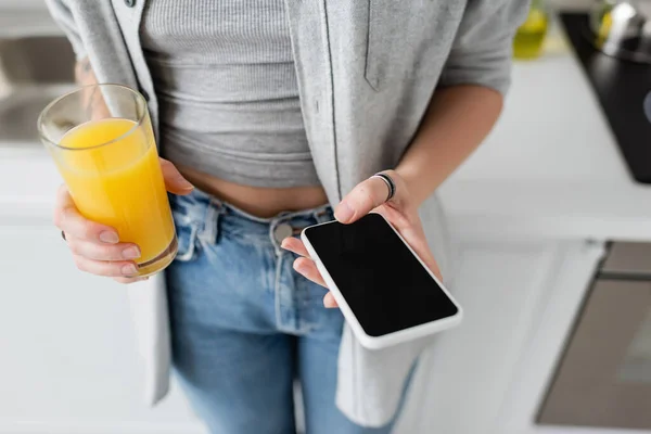 Обрізаний вид молодої жінки з татуюванням на руці, що тримає склянку свіжого апельсинового соку та смартфон з порожнім екраном, стоячи в повсякденному одязі з блакитними джинсами в сучасній квартирі — стокове фото