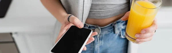Cropped view of young woman with tattoo on hand holding glass of fresh orange juice and smartphone with blank screen while standing in casual clothes with blue denim jeans in modern apartment, banner — Stock Photo