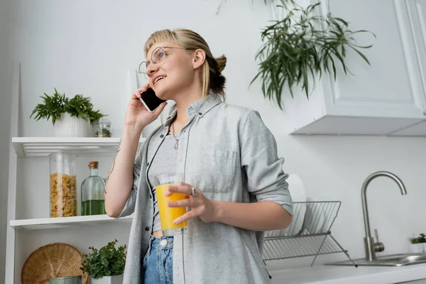 Mujer joven feliz con flequillo y anteojos sosteniendo un vaso de jugo de naranja y hablando en el teléfono inteligente, de pie cerca de las plantas verdes borrosas y estante en la cocina moderna y blanca - foto de stock